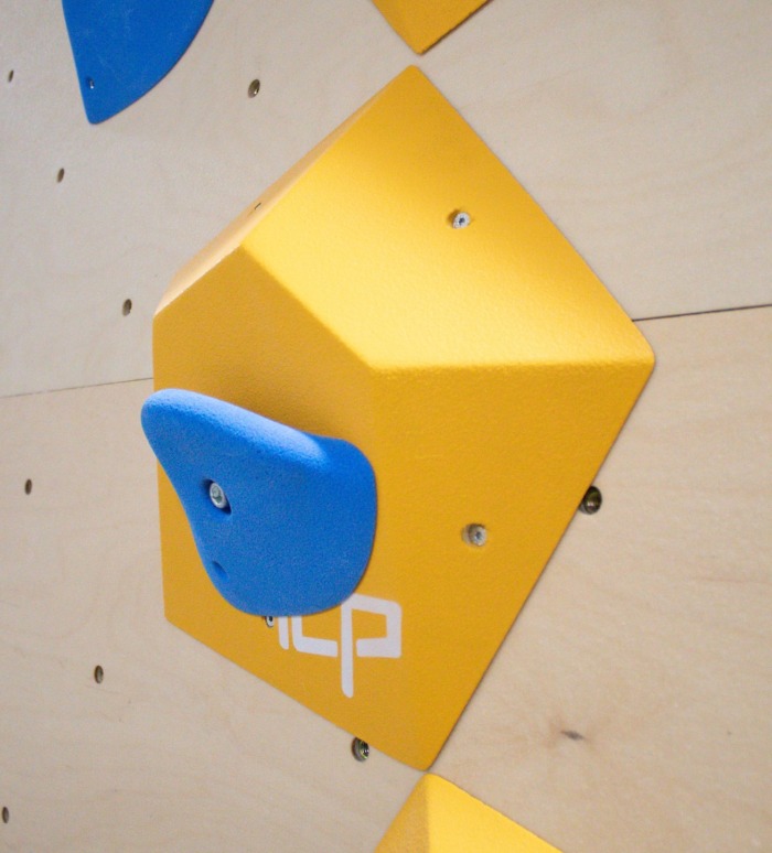 Small yellow ICP Ian I1.3 XS climbing volume on a wood-textured wall, fitted with a blue climbing hold for added route complexity.