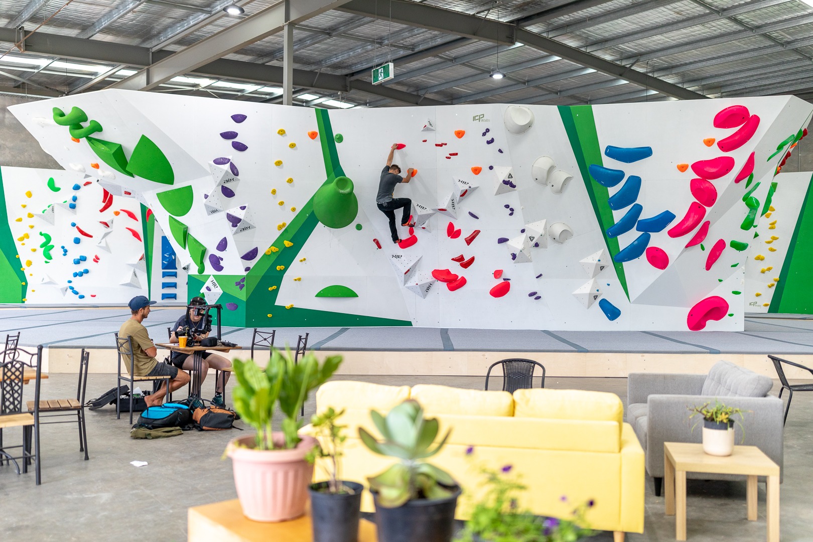 A bouldering gym built by ICP walls, filled with colourful climbing holds. A climber is climbing the holds in the background, while a couple is sitting at tables in the foreground. 