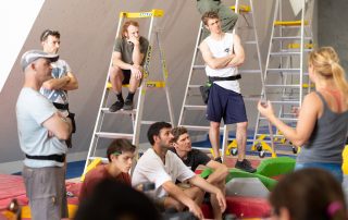 Photo of a route setting workshop with a world cup instructor teaching students seated on ladders and safety mats below an ICP climbing wall.