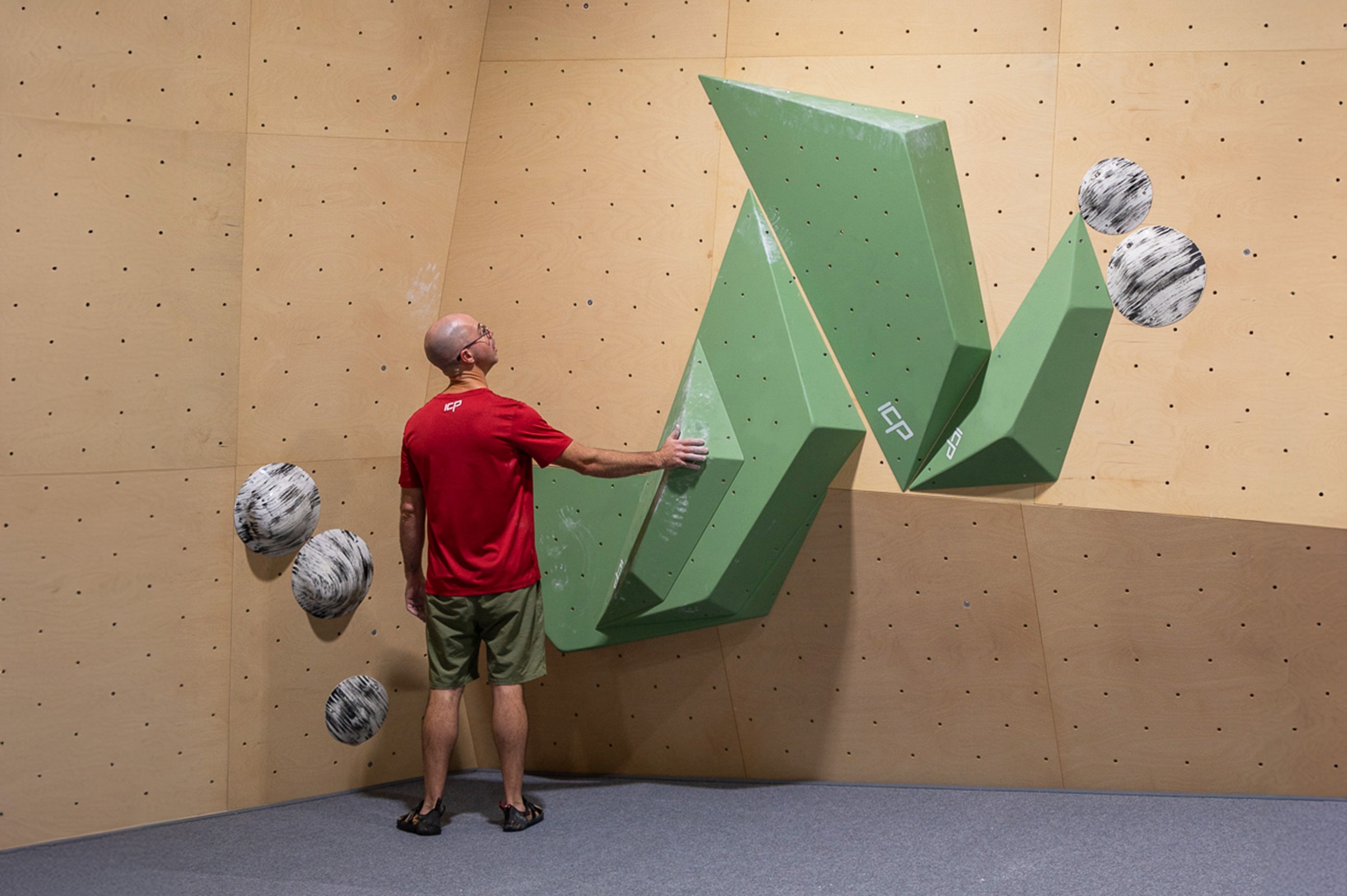 A route setter placing a black hold cross a wooden ICP volume and a climbing wall at ICP Boulderhall & Showroom. The climbing wall is tall and white, which also has a unique wooden detail across the bottom. 