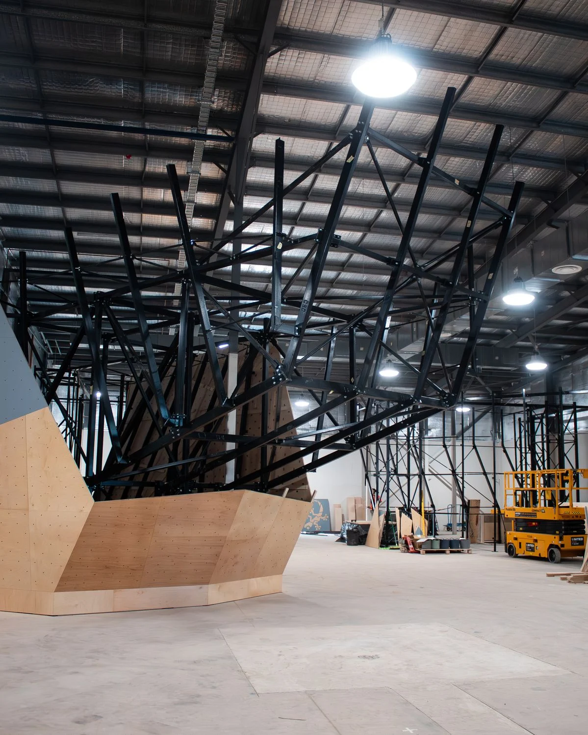 Large indoor construction space featuring a black metal geometric framework over a wooden angular platform; a yellow forklift and scaffolding visible in the background.